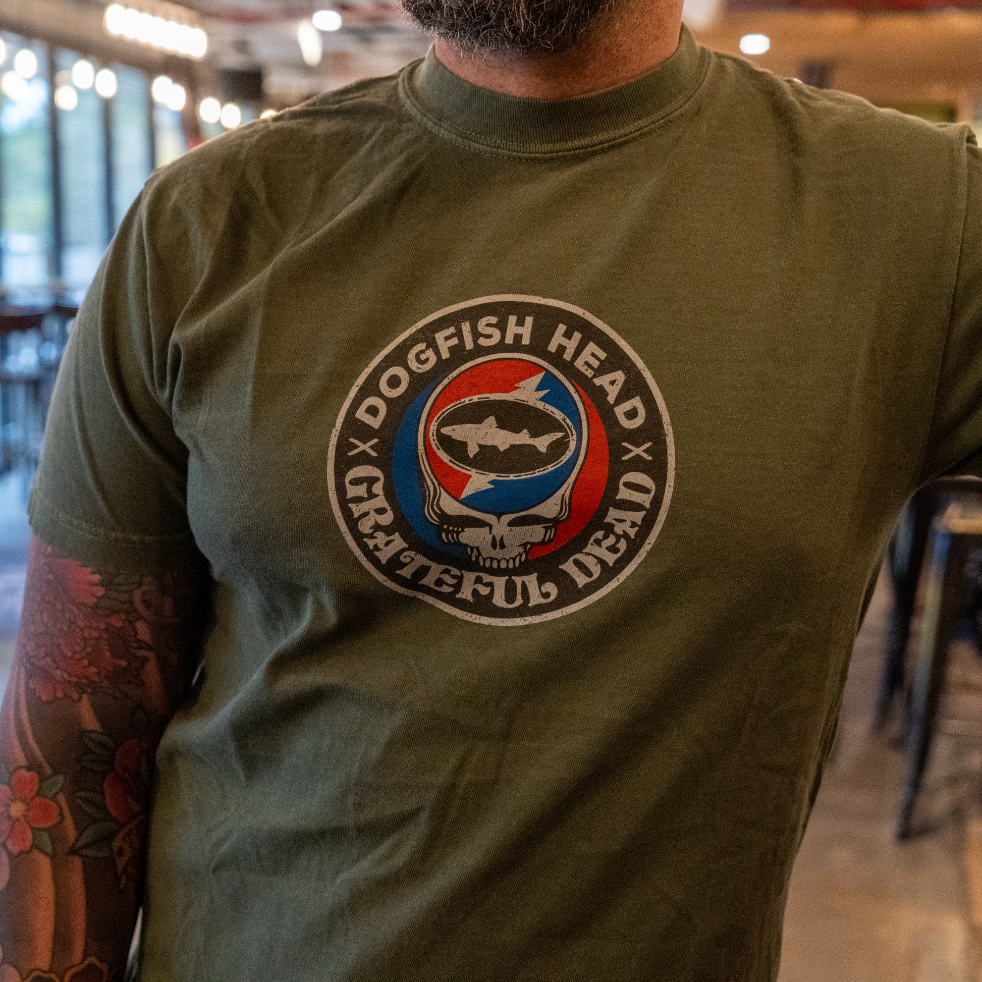 Wearing the Dogfish Head x Grateful Dead Green Logo Tee by GO WEST T-SHIRT COMPANY, a person with colorful tattoos sports the soft cotton shirt featuring a stylized skull, lightning bolt, and fish logo for a unique look.
