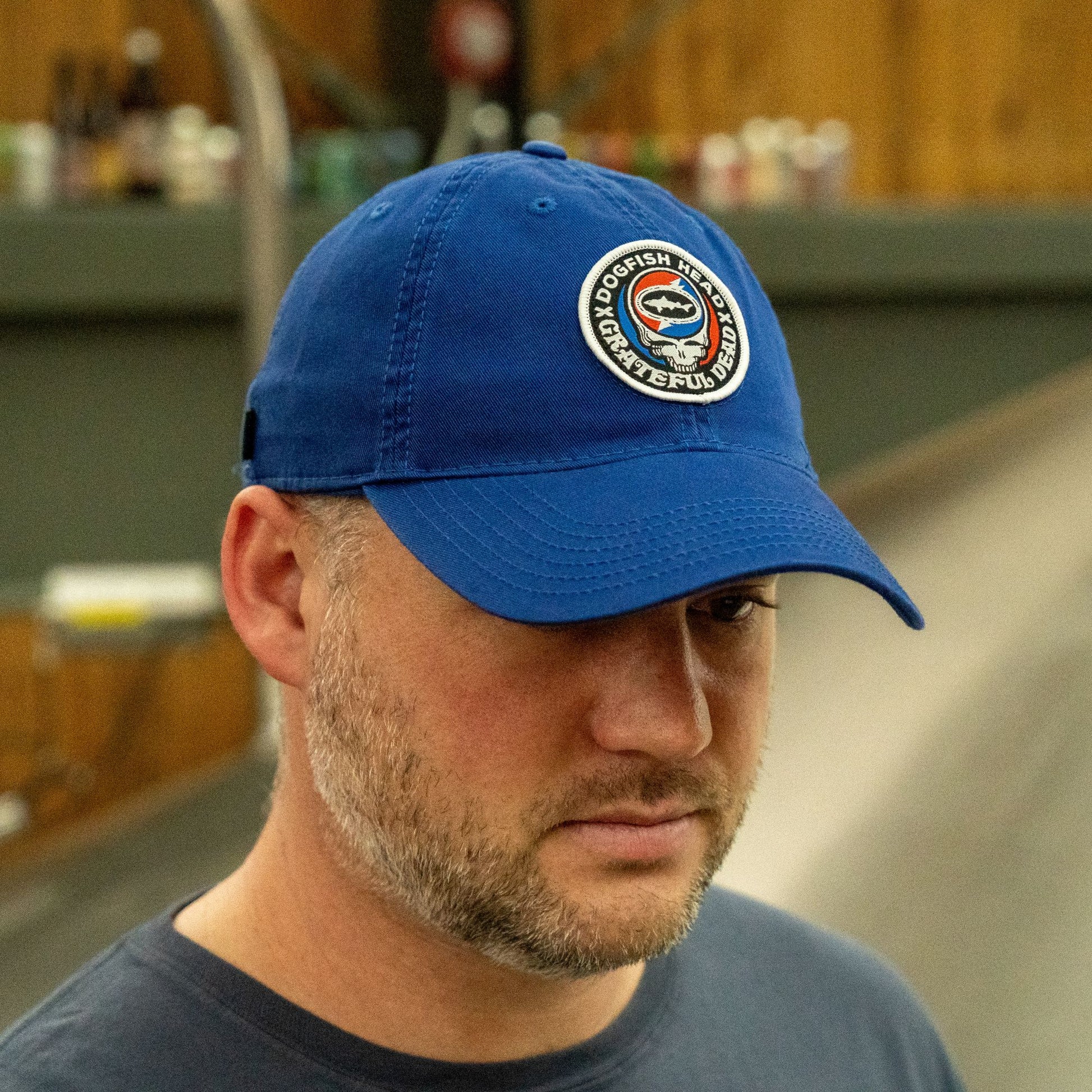 A man with a short beard wears the Dogfish Head x Grateful Dead LEGACY Royal Dad Hat by L2 Brands—an ideal gift for beer lovers—standing indoors with blurred shelves and wooden surfaces in the background.