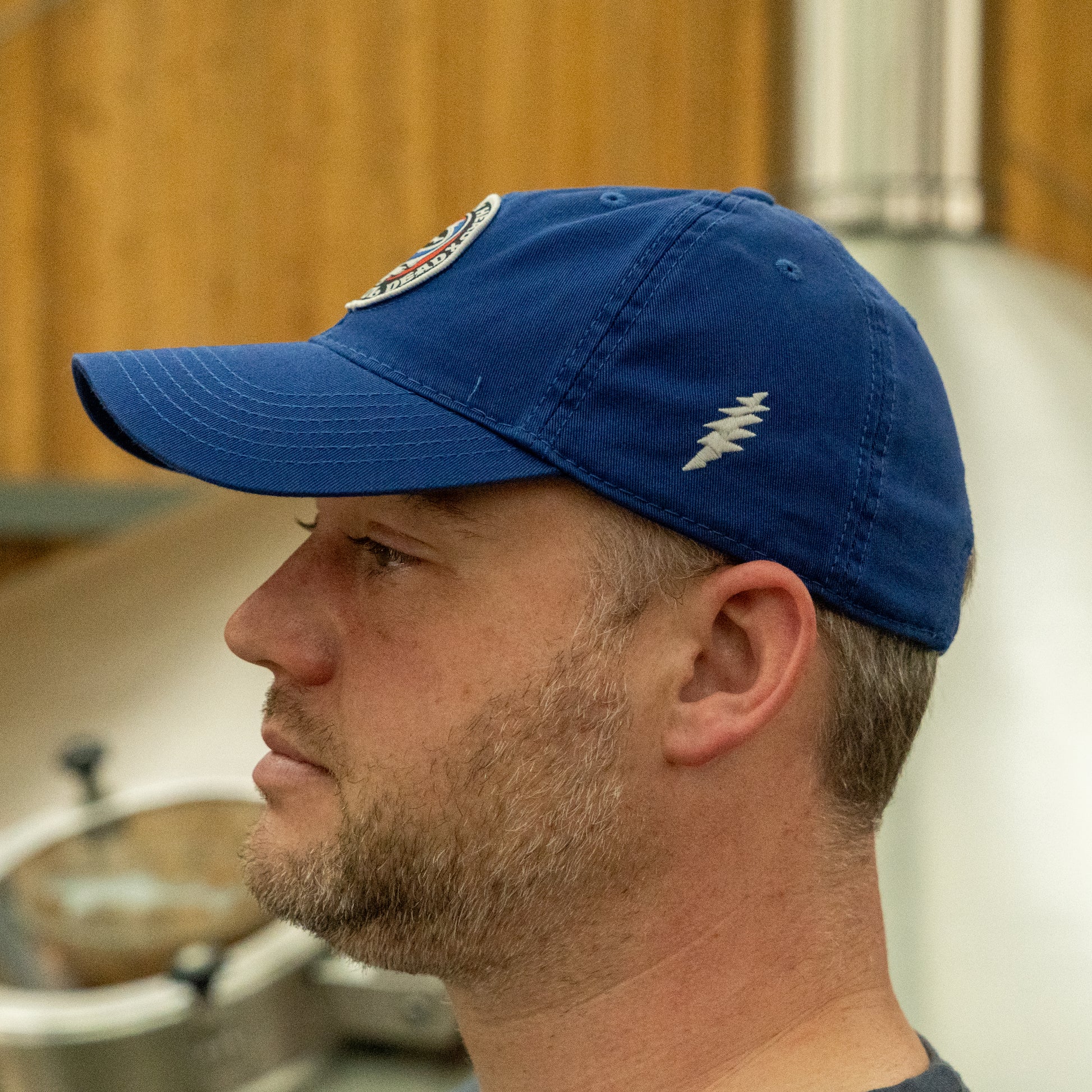 A man with a short beard, seen in profile, wears the L2 Brands Dogfish Head x Grateful Dead LEGACY Royal Dad Hat. Behind him is a wooden wall and metal equipment, evoking an indoor brewery — great inspiration for beer lovers’ gifts.