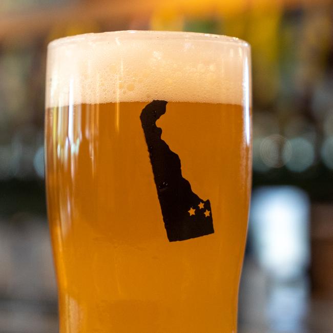 The Dogfish Head Brewhouse Glass by Sahm Glass Inc showcases Delaware pride with its black outline of the state and three stars, filled with amber beer against a blurred background.