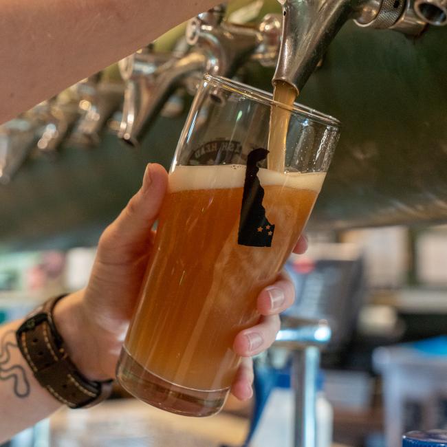 A hand holds a Dogfish Head Brewhouse Glass by Sahm Glass Inc under a beer tap, filling with hazy amber beer as foam forms on top. Multiple taps in the background evoke true Delaware pride.