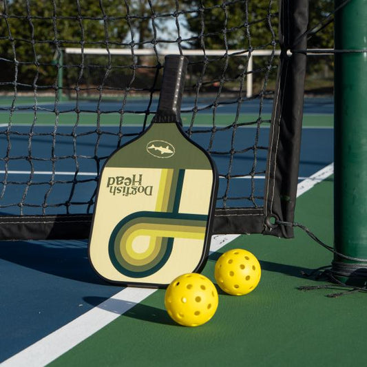 The Dogfish Head Pickleball Paddle by Custom Pickleball - MAH Tech leans against a tennis net on an outdoor court, with three yellow pickleballs nearby.