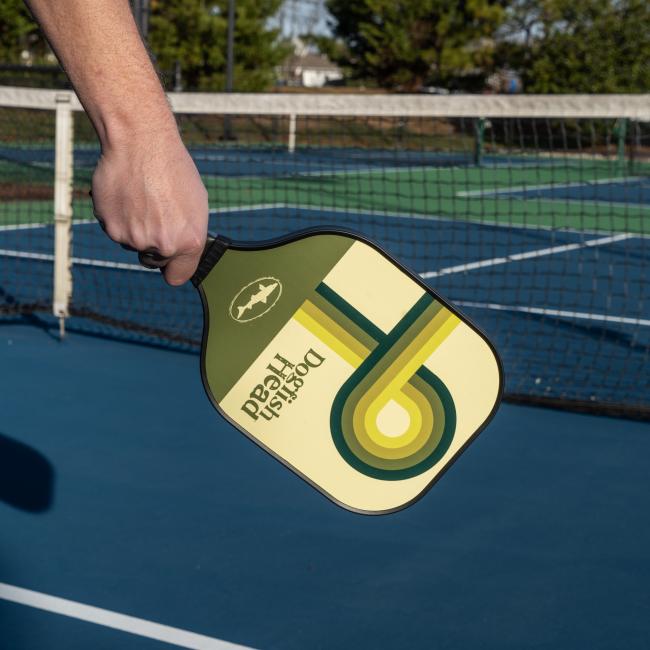 A person holds the Dogfish Head Pickleball Paddle by Custom Pickleball - MAH Tech, featuring a honeycomb core, on a blue outdoor court near the net.