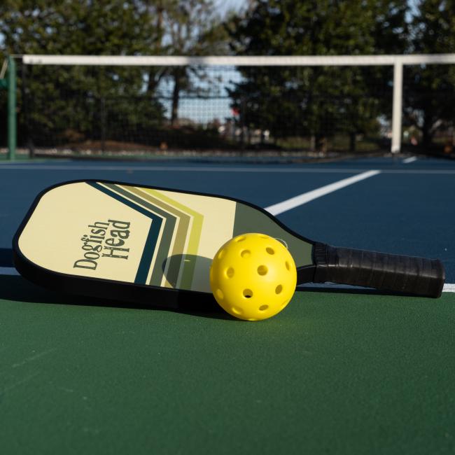 A Dogfish Head Pickleball Paddle by Custom Pickleball - MAH Tech and a yellow perforated ball rest on an outdoor court near the net, with trees visible in the background.