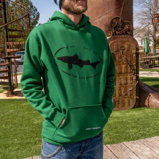 A man in a Dogfish Head x Boathouse Green Heavyweight Hoodie by Boathouse Apparel stands outside by a tall, rusted metal structure and artificial grass, wearing a beanie and sunglasses.