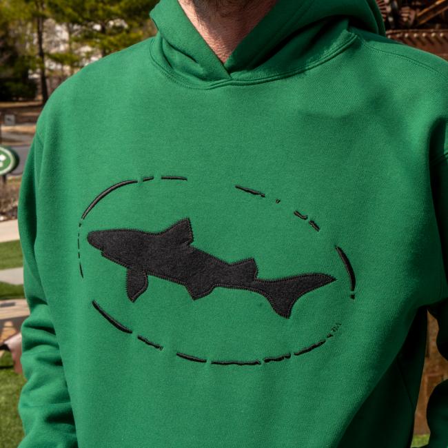 A person wears a Dogfish Head x Boathouse Green Heavyweight Hoodie by Boathouse Apparel, showing only the upper torso and featuring a large black shark silhouette inside a dashed oval on the front.