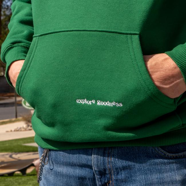 A person stands outdoors in sunlight wearing blue jeans and a green Dogfish Head x Boathouse Heavyweight Hoodie from Boathouse Apparel, featuring "explore goodness" embroidered in white above the pocket.