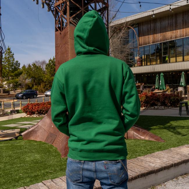 Wearing a Dogfish Head x Boathouse Green Heavyweight Hoodie by Boathouse Apparel and blue jeans, a person stands outdoors with their back to the camera, near a large metal sculpture and a modern building with outdoor seating.