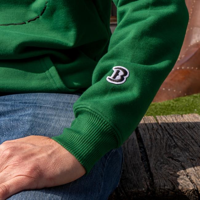 A person wearing the Boathouse Apparel Dogfish Head x Boathouse Green Heavyweight Hoodie with a black and white letter B patch on the sleeve sits on a wooden bench, showing part of their arm and blue jeans.