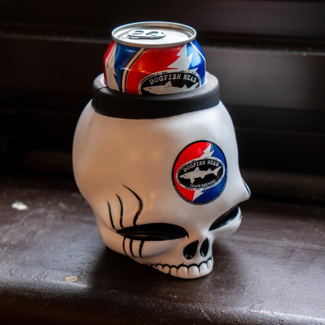The Bottoms Up Dogfish Head x Grateful Dead 3D Can Cooler, shaped like a white skull with black eye sockets, holds a Dogfish Head beer can featuring red, white, and blue logos—perfect for fans of both brands and the Grateful Dead.