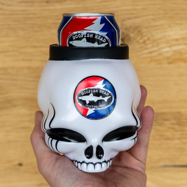 A hand holds a white skull-shaped Dogfish Head x Grateful Dead 3D Can Cooler by Bottoms Up, with black details and a Dogfish Head beer can inside, featuring the brand’s logo sticker on the front, set on a light wood surface.
