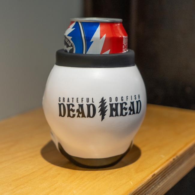 A can with a red, blue, and white design sits in a Bottoms Up Dogfish Head x Grateful Dead 3D Can Cooler on a wooden surface.