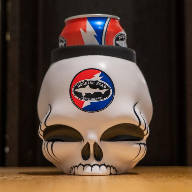 A can of Dogfish Head beer rests in the Bottoms Up Dogfish Head x Grateful Dead 3D Can Cooler, shaped like a white skull with the brewery’s logo—a striking tribute to Grateful Dead fans. The dark background is out of focus.