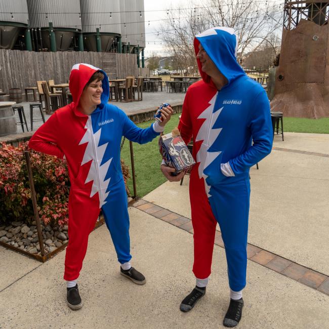 Two people in matching Dogfish Head x Grateful Dead Adult Onesies by In Your Face Apparel stand outside; one holds a microphone, the other a box. Both are smiling, sharing a lighthearted moment with serious Grateful Dead vibes.