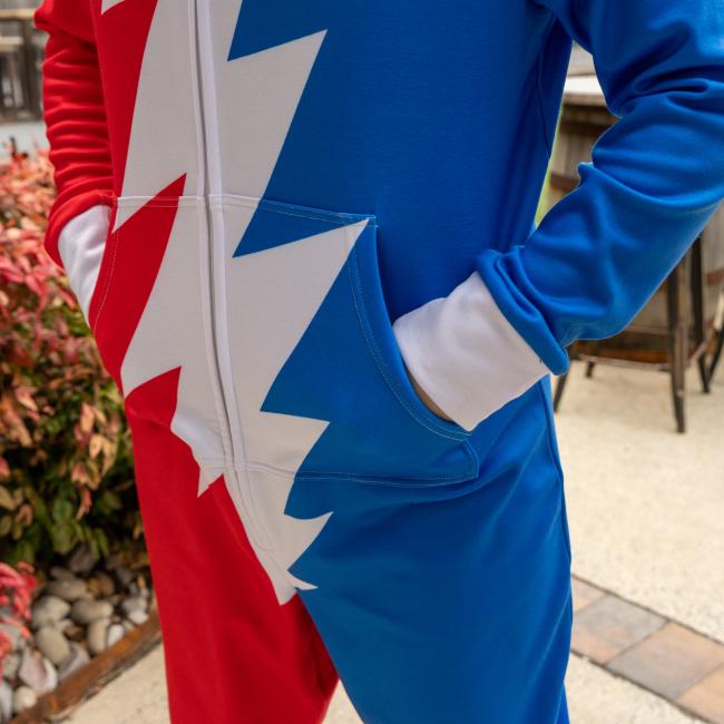 A person wearing the Dogfish Head x Grateful Dead Adult Onesie by In Your Face Apparel, featuring a blue and red design with a white jagged stripe, stands outdoors with hands in pockets near plants and patio furniture.
