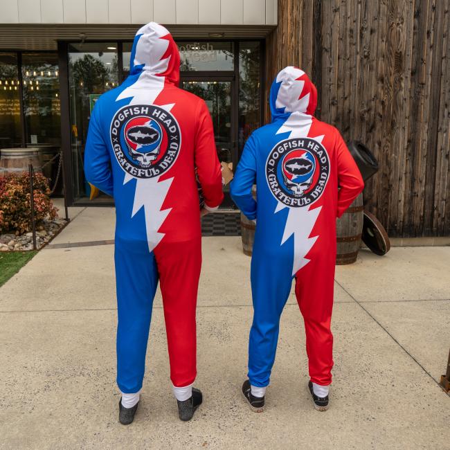 Two people, backs to the camera, stand by a rustic building in red, white, and blue Dogfish Head x Grateful Dead Adult Onesies from In Your Face Apparel featuring lightning bolts and a circular logo on the back.