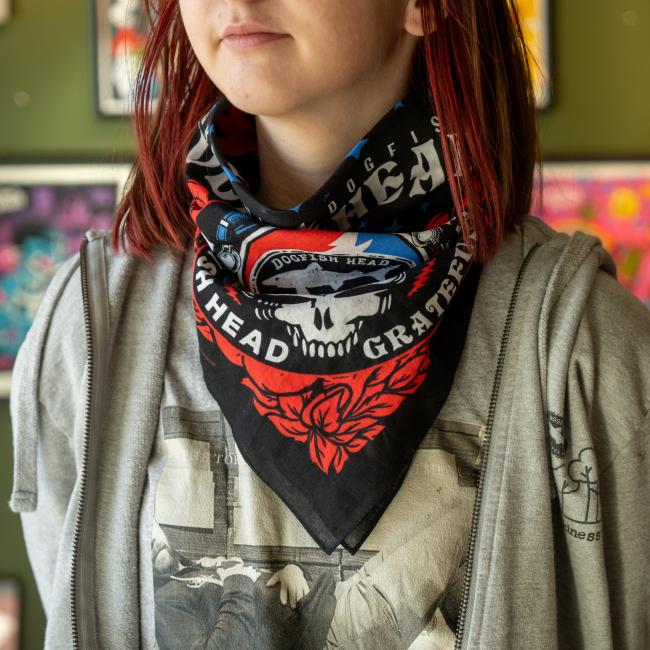 A person with shoulder-length reddish hair wears a Merchtank Dogfish Head x Grateful Dead Bandana, featuring a skull and rose design, along with a gray zip-up hoodie and graphic tee. Colorful art is visible in the blurred background.