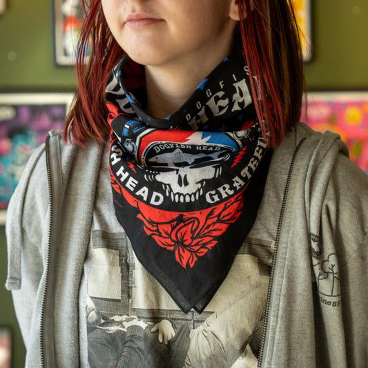 A person with red-streaked hair wears a Merchtank Dogfish Head x Grateful Dead Black Bandana featuring a skull design over a gray graphic T-shirt and light gray hoodie. Colorful posters appear in the blurred background.