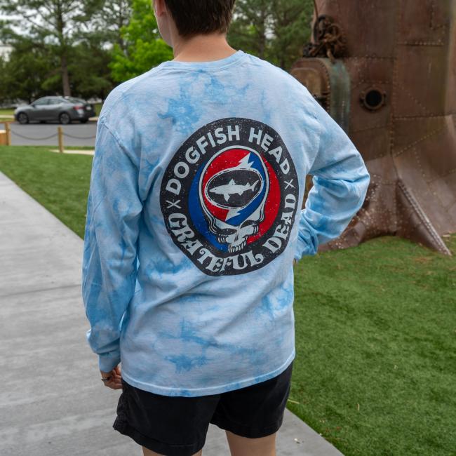 Someone stands on a sidewalk by grass and trees, wearing the GO WEST T-SHIRT COMPANY Dogfish Head x Grateful Dead Blue Tie Dye Long Sleeve Tee, featuring a large graphic on the back.