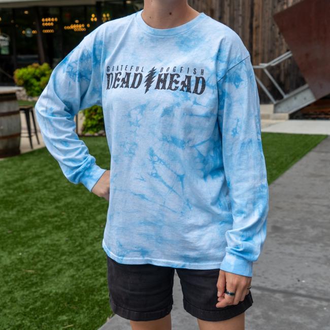 A person stands outdoors on artificial grass, wearing a GO WEST T-SHIRT COMPANY Dogfish Head x Grateful Dead Blue Tie Dye Long Sleeve Tee and black shorts. Their face is not visible.