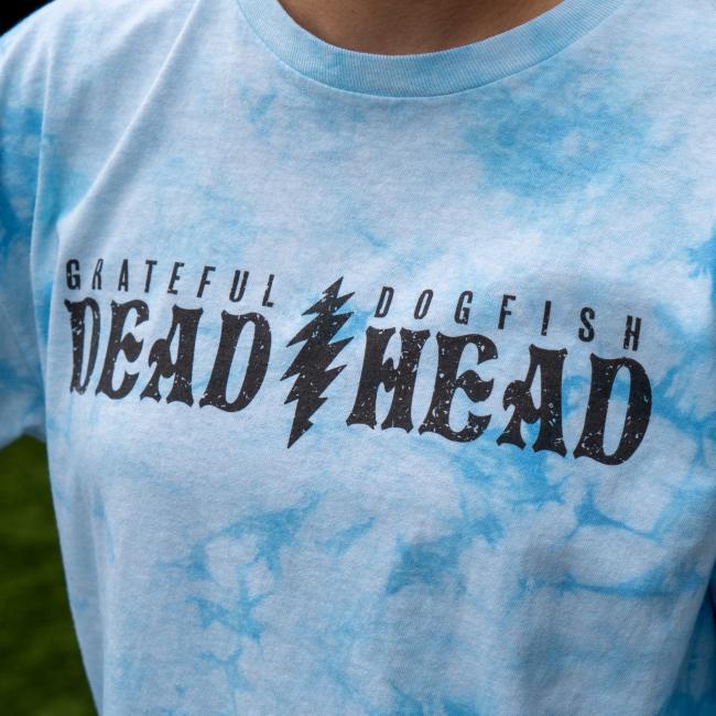 A person wears the Dogfish Head x Grateful Dead Blue Tie Dye Long Sleeve Tee by GO WEST T-SHIRT COMPANY, featuring "GRATEFUL DOGFISH DEAD HEAD" text and a lightning bolt graphic inspired by Grateful Dead style.