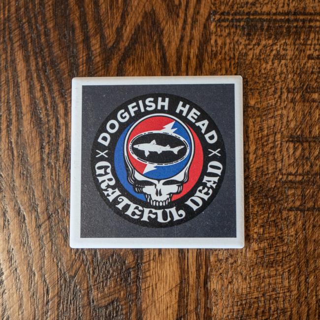 The Dogfish Head x Grateful Dead Ceramic Coaster by Paint the Town, Inc. features a fish inside a skull with a red and blue lightning bolt background, displayed on a wooden surface.