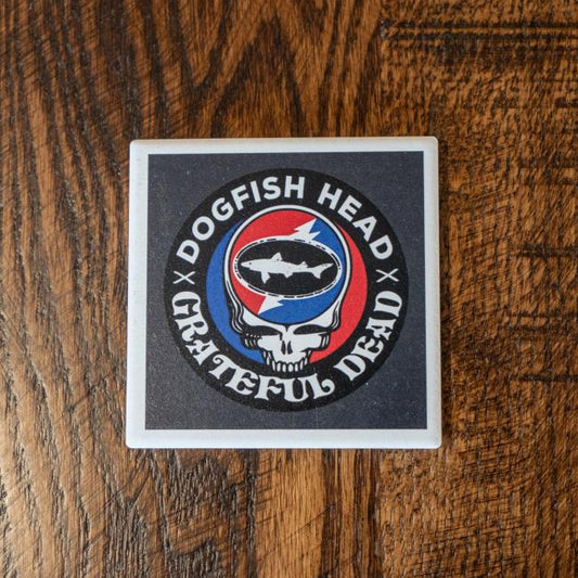 The Dogfish Head x Grateful Dead Ceramic Coaster by Paint the Town, Inc. features a fish inside a skull with a red and blue lightning bolt background, displayed on a wooden surface.