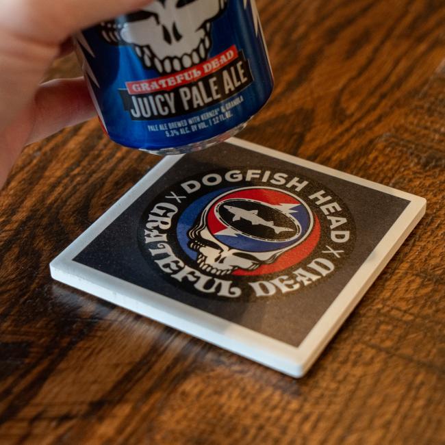 A hand holds a can above a Paint the Town, Inc. Dogfish Head x Grateful Dead Ceramic Coaster, which features both logos, on a wooden surface.