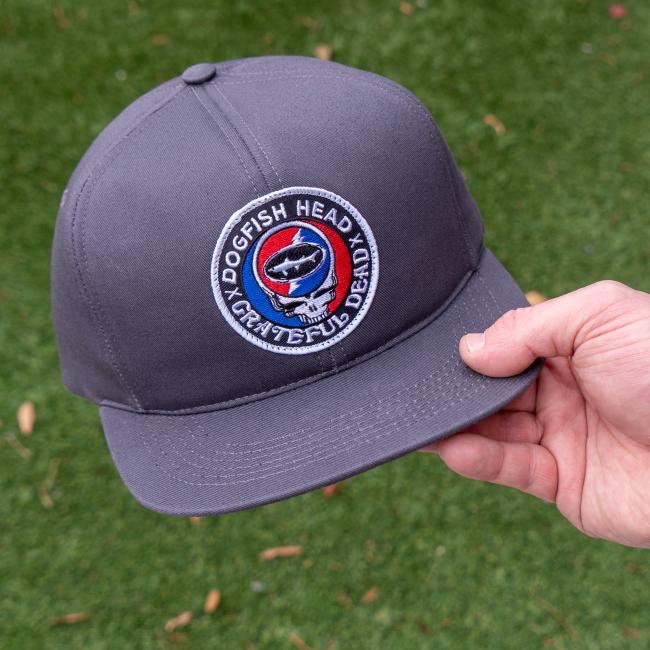 A hand holds the Dogfish Head x Grateful Dead Charcoal Flat Brim by GRAFFITI INC, featuring a round patch with a shark inside a red, white, and blue Steal Your Face logo. The vintage snapback is set against green grass.