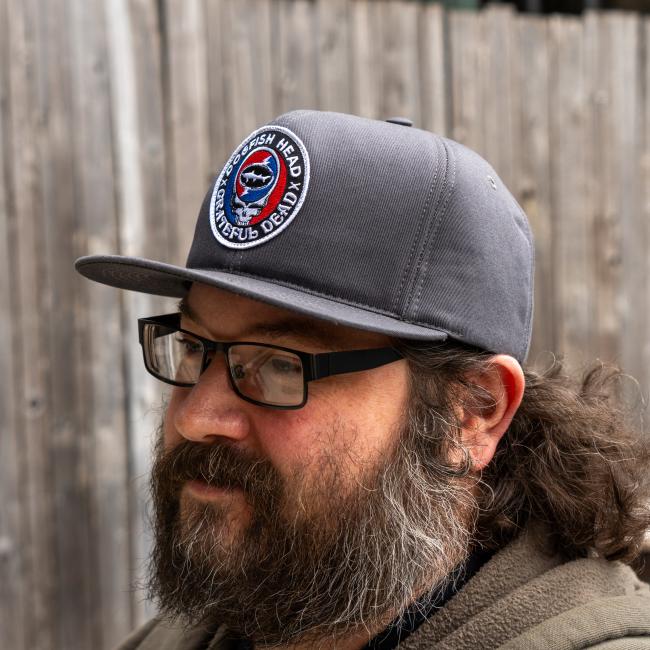 A bearded man with glasses wears the GRAFFITI INC Dogfish Head x Grateful Dead Charcoal Flat Brim hat while standing by a wooden fence.