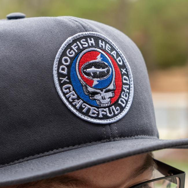 A close-up of the Dogfish Head x Grateful Dead Charcoal Flat Brim by GRAFFITI INC, featuring a round patch with skull, fish silhouette, and red-blue background—a must-have vintage snapback for fans.