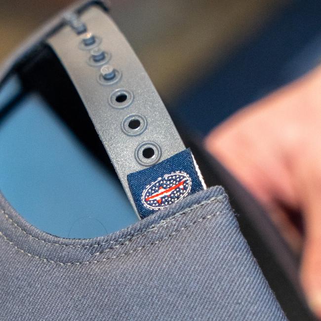 Close-up of the Dogfish Head x Grateful Dead Charcoal Flat Brim by GRAFFITI INC, showing its adjustable plastic strap, fabric tag with a red, white, and blue oval logo featuring stars and stripes, and a hand holding the hat.