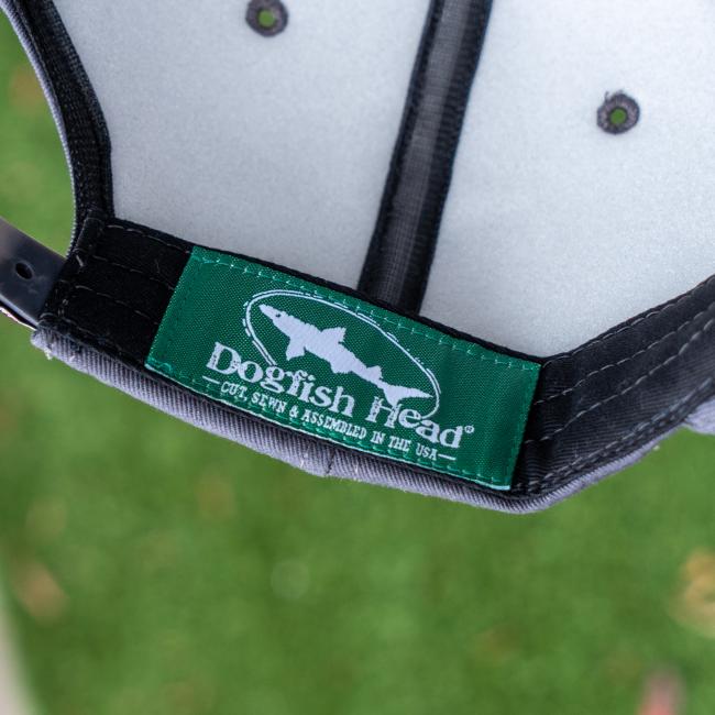 Close-up of the inside of a Dogfish Head x Grateful Dead Charcoal Flat Brim by GRAFFITI INC, showing a green label with fish logo and "Dogfish Head® Cut, Sewn & Assembled in the USA," set against blurred green grass for a vintage snapback feel.