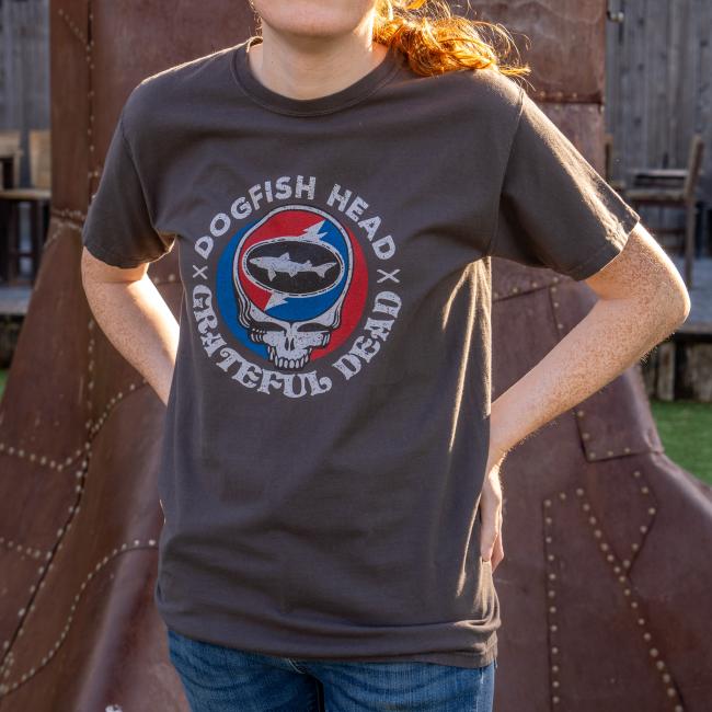 A person stands outdoors, hands on hips, wearing a Dogfish Head x Grateful Dead Dark Grey Short Sleeve Tee by GO WEST T-SHIRT COMPANY. Their face is partially out of frame.