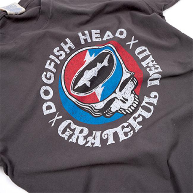 The GO WEST T-SHIRT COMPANY Dogfish Head x Grateful Dead Dark Grey Short Sleeve Tee features both iconic logos, a skull graphic with a red and blue backdrop, and bold white text—perfect for craft beer and music fans.