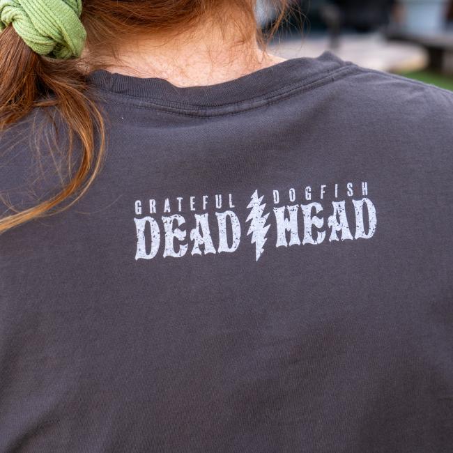 A person with brown hair in a green scrunchie wears a GO WEST T-SHIRT COMPANY Dogfish Head x Grateful Dead dark grey short sleeve tee with white text on the back that reads: Grateful Dogfish Dead Head.