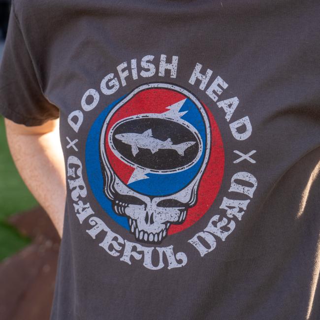 A person wears the Dogfish Head x Grateful Dead Dark Grey Short Sleeve Tee by GO WEST T-SHIRT COMPANY, featuring a skull, shark in an oval, and red and blue split background—ideal for craft beer t-shirt collectors.