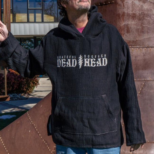 A person wearing a black Dogfish Head x Grateful Dead Fiber Art Hoodie by Pure Country Inc - Fiber Art stands outdoors near a large rust-colored metal structure, with their face partially out of frame.