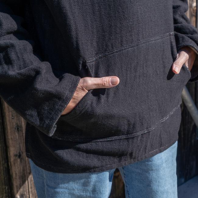 A person wearing the Dogfish Head x Grateful Dead Fiber Art Hoodie by Pure Country Inc - Fiber Art stands with hands in the front pocket, set against a wooden background.