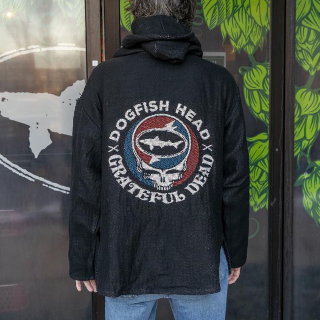 A person stands with their back to the camera, wearing the Pure Country Inc - Fiber Art Dogfish Head x Grateful Dead Fiber Art Hoodie featuring a circular shark and skull logo. Green leaves and a white whale logo are visible in the background.
