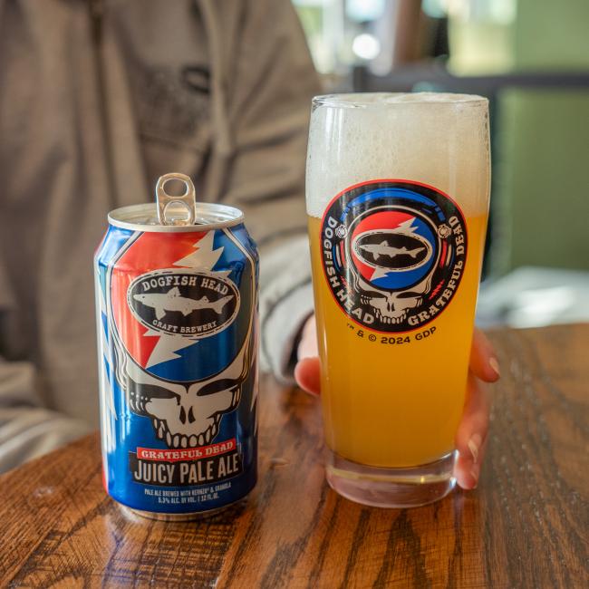 A person holds a Dogfish Head x Grateful Dead Glass by Grandstand Glassware & Apparel filled with hazy pale ale next to an open can on a wooden table. Both feature vibrant Grateful Dead-themed artwork.