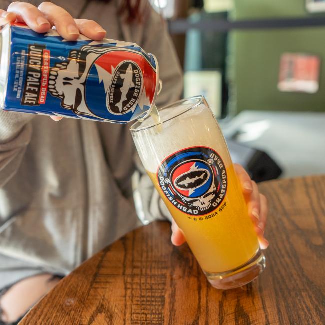 A person pours Dogfish Head Juicy Pale Ale into a Dogfish Head x Grateful Dead Glass by Grandstand Glassware & Apparel, forming a foamy head atop the beer at an indoor wooden table.