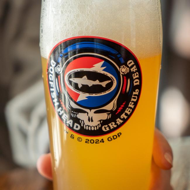 A hand holds the hazy yellow Dogfish Head x Grateful Dead Glass by Grandstand Glassware & Apparel, featuring a colorful logo with a shark silhouette and stylized skull.