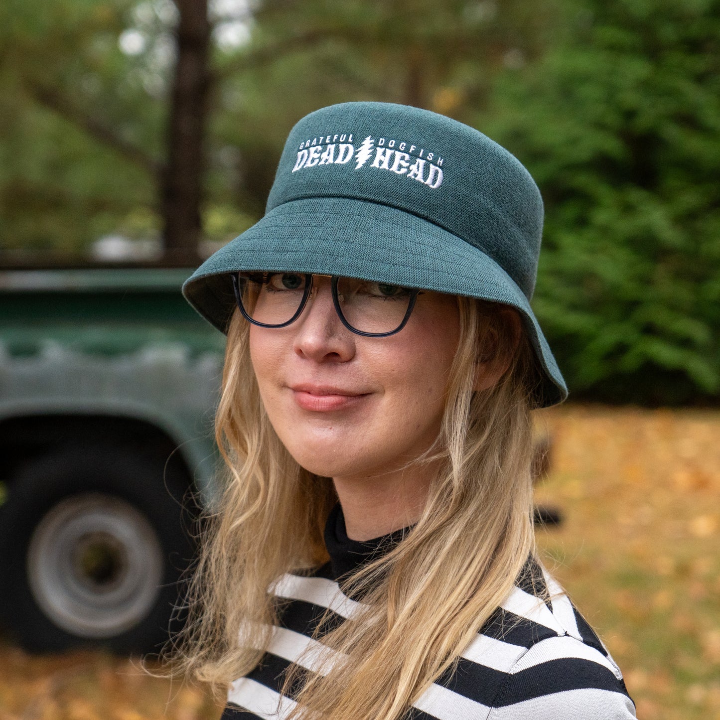 A person with long blonde hair, glasses, and a dark green Dogfish Head x Grateful Dead KANGOL Bucket Hat by BOLLMAN HATS stands outdoors, smiling in a black-and-white striped shirt, with blurred trees and a vehicle behind them.
