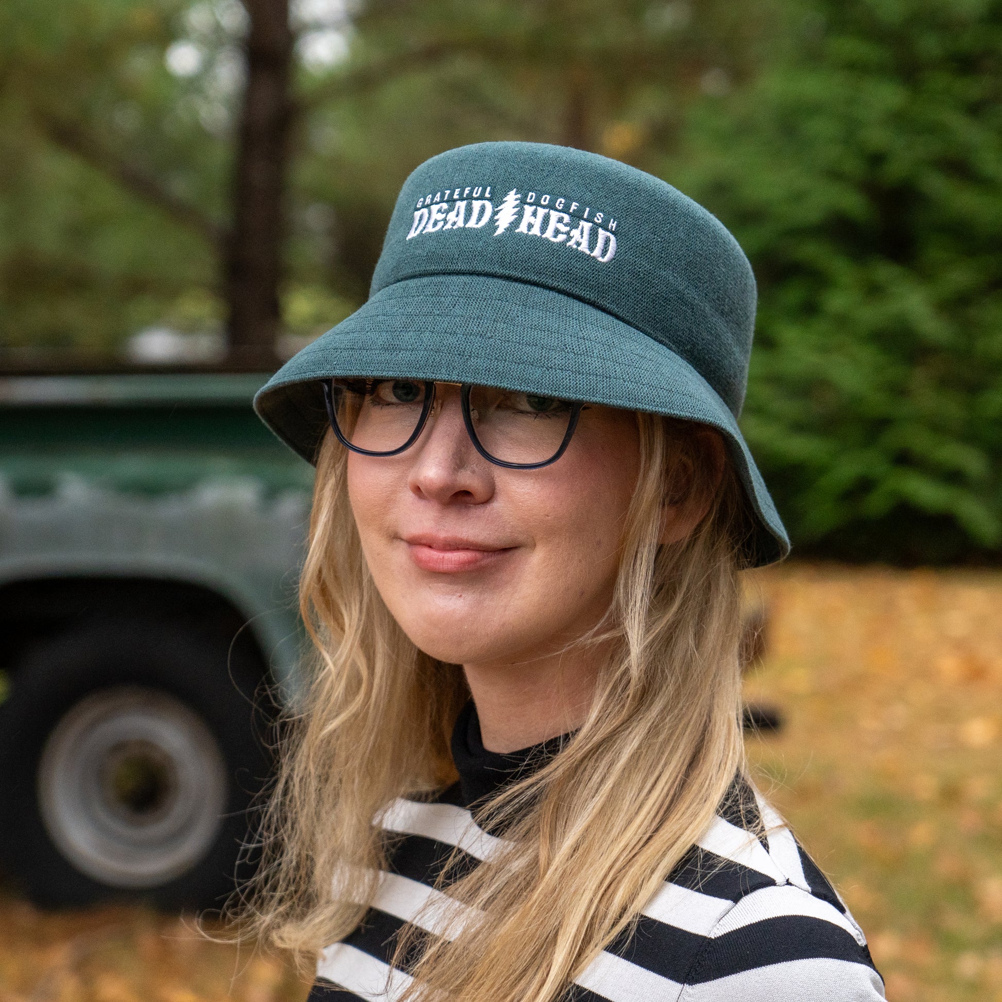 A person with long blonde hair, glasses, and a dark green Dogfish Head x Grateful Dead KANGOL Bucket Hat by BOLLMAN HATS stands outdoors, smiling in a black-and-white striped shirt, with blurred trees and a vehicle behind them.