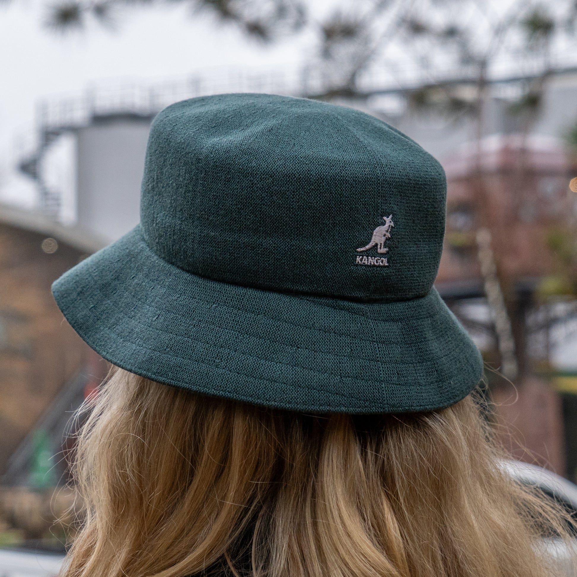 A person with long blonde hair is seen from behind wearing a dark green Dogfish Head x Grateful Dead KANGOL Bucket Hat by BOLLMAN HATS outdoors, with blurred buildings and trees in the background.