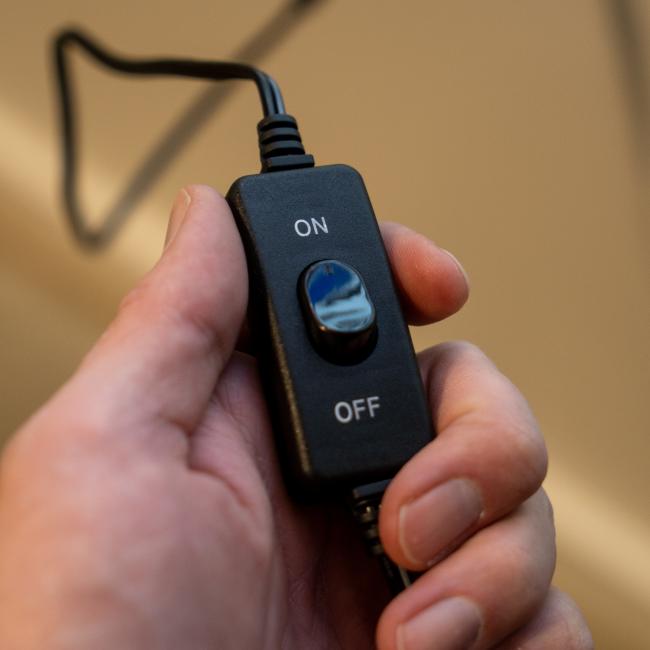 A hand holds a black inline switch with a blue ON/OFF button, attached to a black cord—ideal for controlling your Firebolt Group Dogfish Head x Grateful Dead LED Sign or other bar decor.