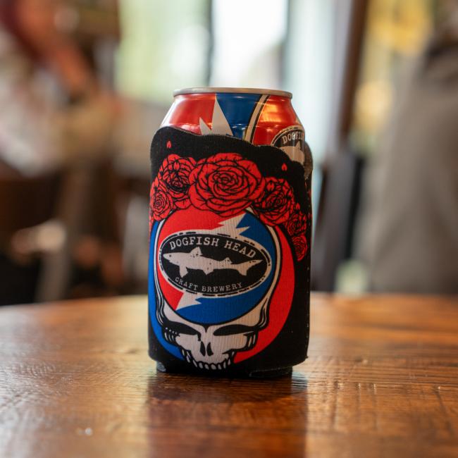 The Grandstand Glassware & Apparel Dogfish Head x Grateful Dead Skull and Roses Can Cooler, featuring the iconic skull, lightning bolt, and red roses design, rests on a wooden table—ideal as a vibrant festival drink accessory.