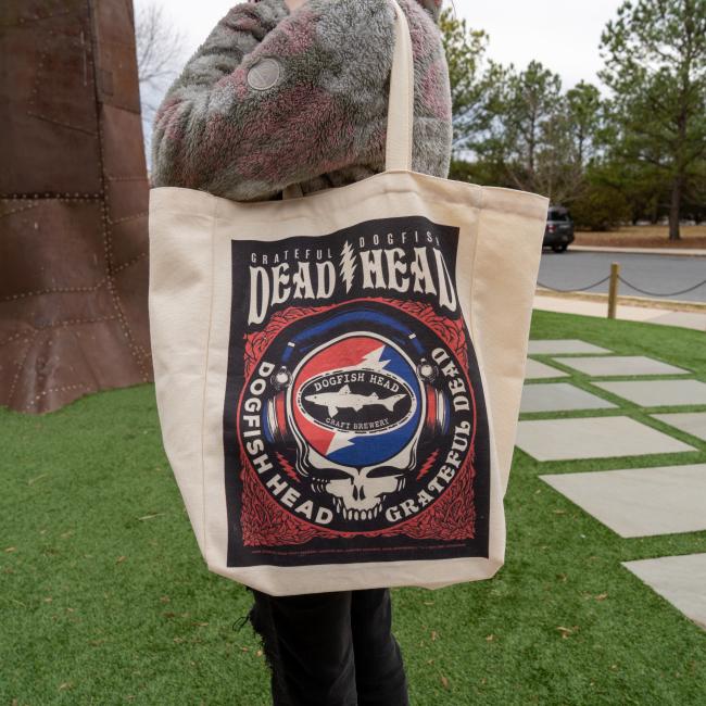 A person holds a beige Dogfish Head x Grateful Dead Tote Bag by Red Promotions, featuring a unique graphic and standing outdoors on green grass—ideal craft beer merchandise for fans.