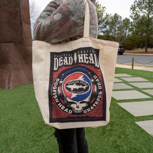 A person holds a beige Dogfish Head x Grateful Dead Tote Bag by Red Promotions, featuring a unique graphic and standing outdoors on green grass—ideal craft beer merchandise for fans.